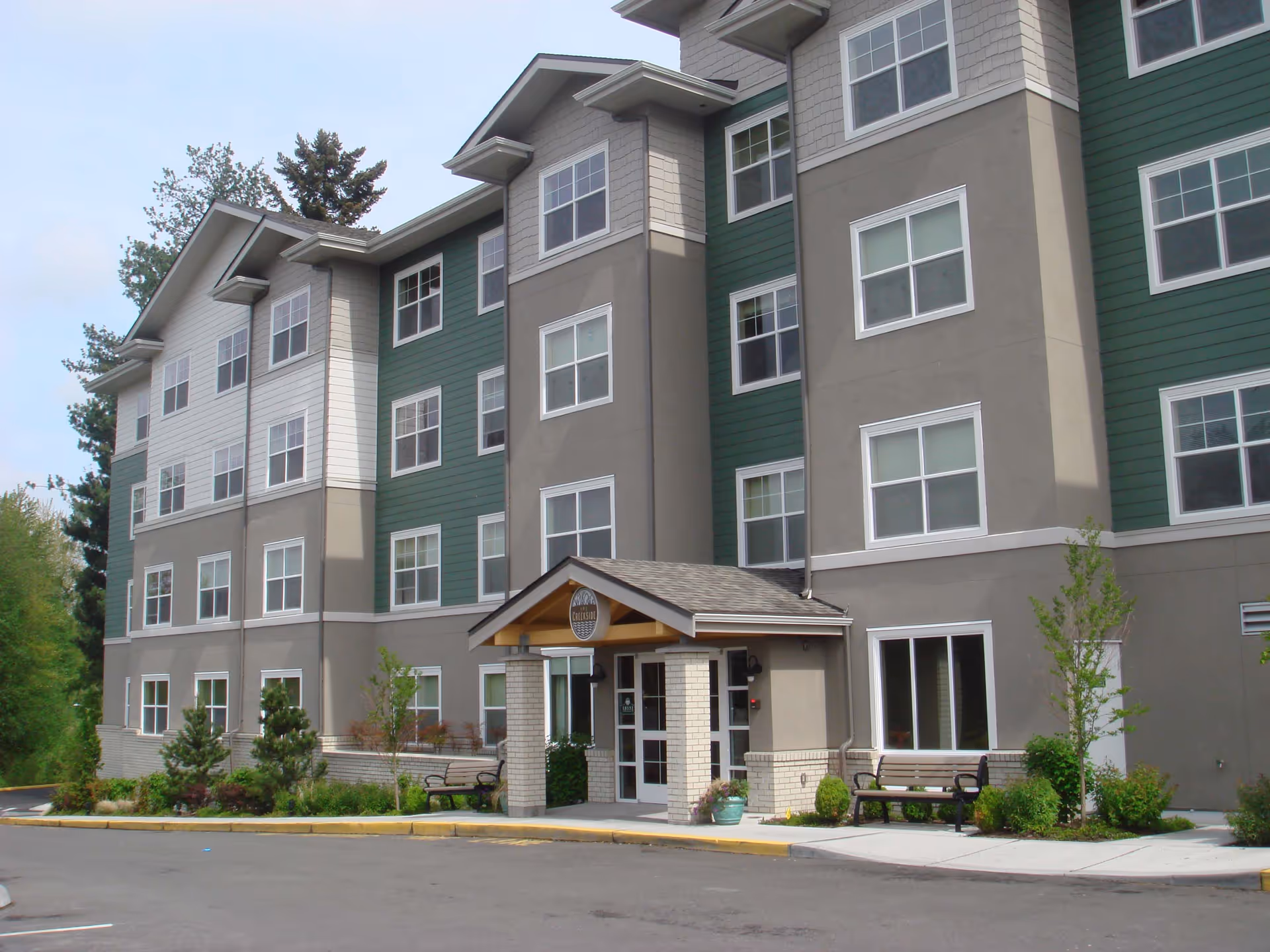 Exterior view of a multi-story senior living facility named The Creekside with a covered entrance, benches, small trees, and shrubs along the front sidewalk.