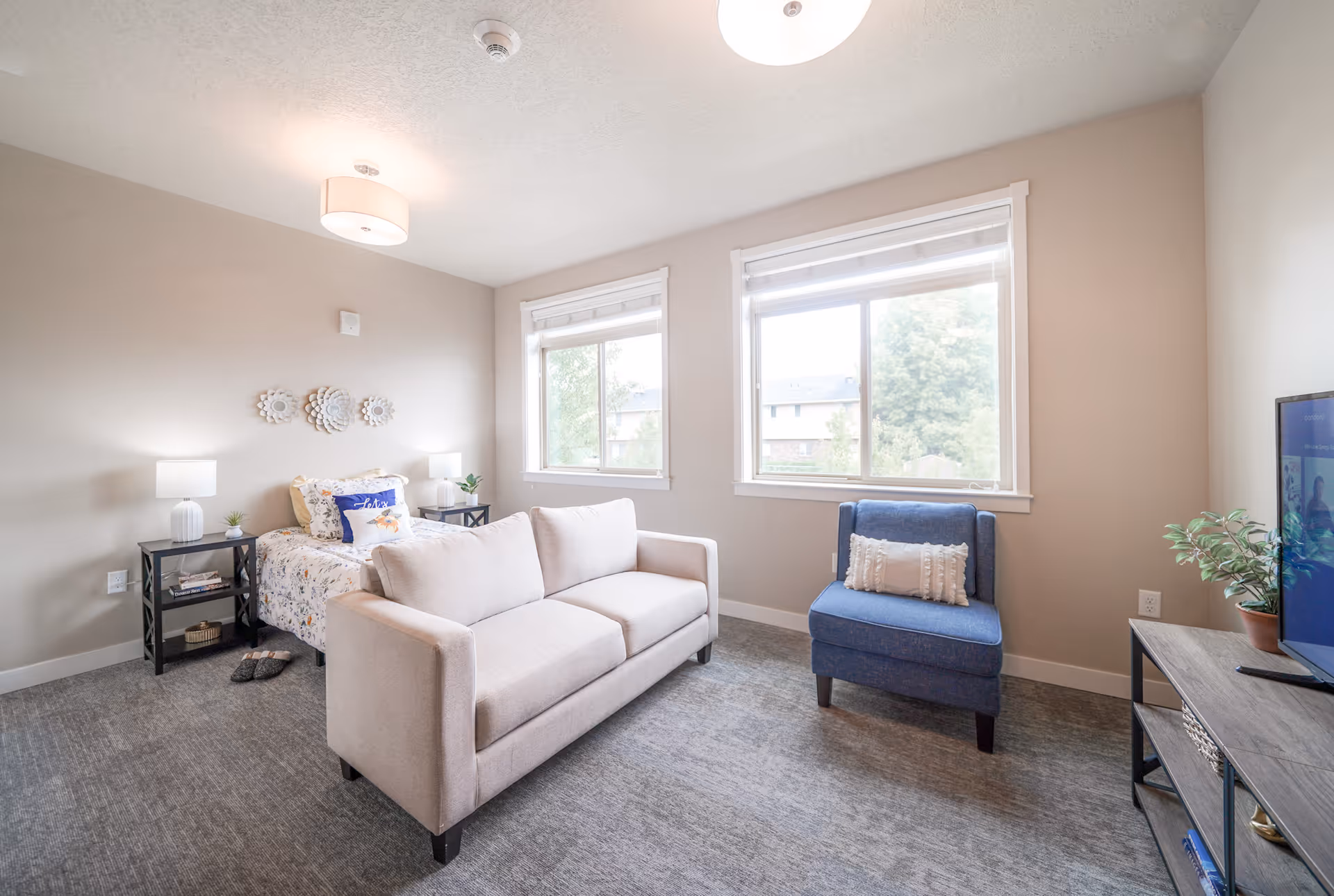 A bright and cozy senior living room featuring a beige sofa, a blue armchair with a decorative pillow, a bed with floral bedding, two bedside tables with lamps, and a TV on a stand. Two large windows let in natural light and show greenery outside.