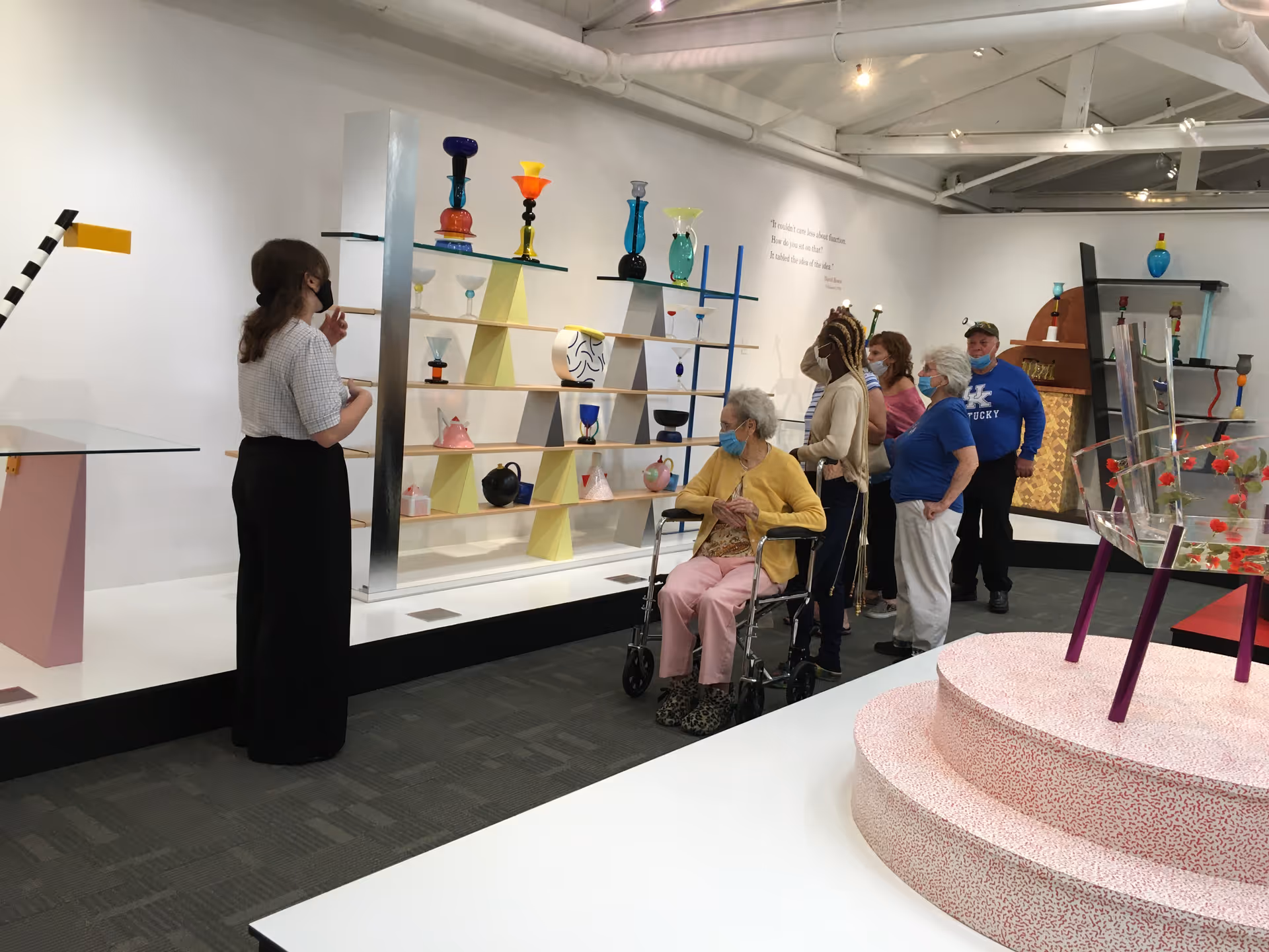 A group of elderly people, some wearing masks, are observing colorful glass art pieces displayed on shelves and stands in a gallery-like setting. One woman in a wheelchair is in the foreground, and a guide or staff member is explaining the exhibits.