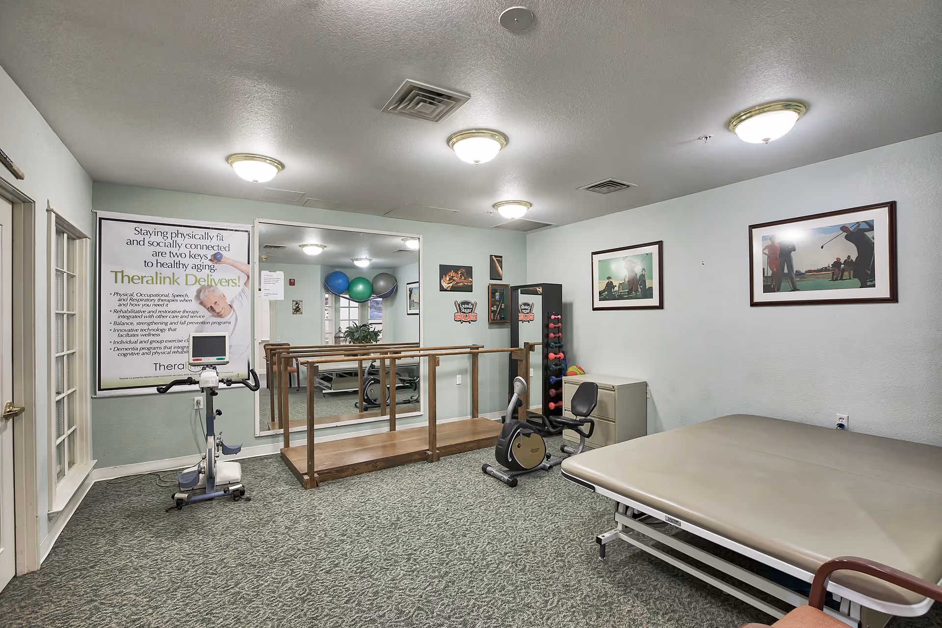 A physical therapy or exercise room with a stationary exercise bike, a wooden balance beam with handrails, a recumbent exercise bike, a rack of colorful dumbbells, and a large padded therapy table. The walls are light green with framed pictures and a large mirror. There is a poster on the wall promoting Theralink services for healthy aging.
