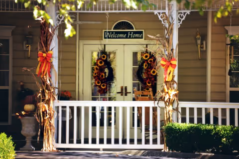 Front porch entrance of a building with double glass doors decorated with sunflower wreaths. A sign above the doors reads 'Welcome Home'. The porch has white railings and two columns wrapped with corn stalks tied with red and yellow bows. There is a rocking chair on the porch and some greenery in front.