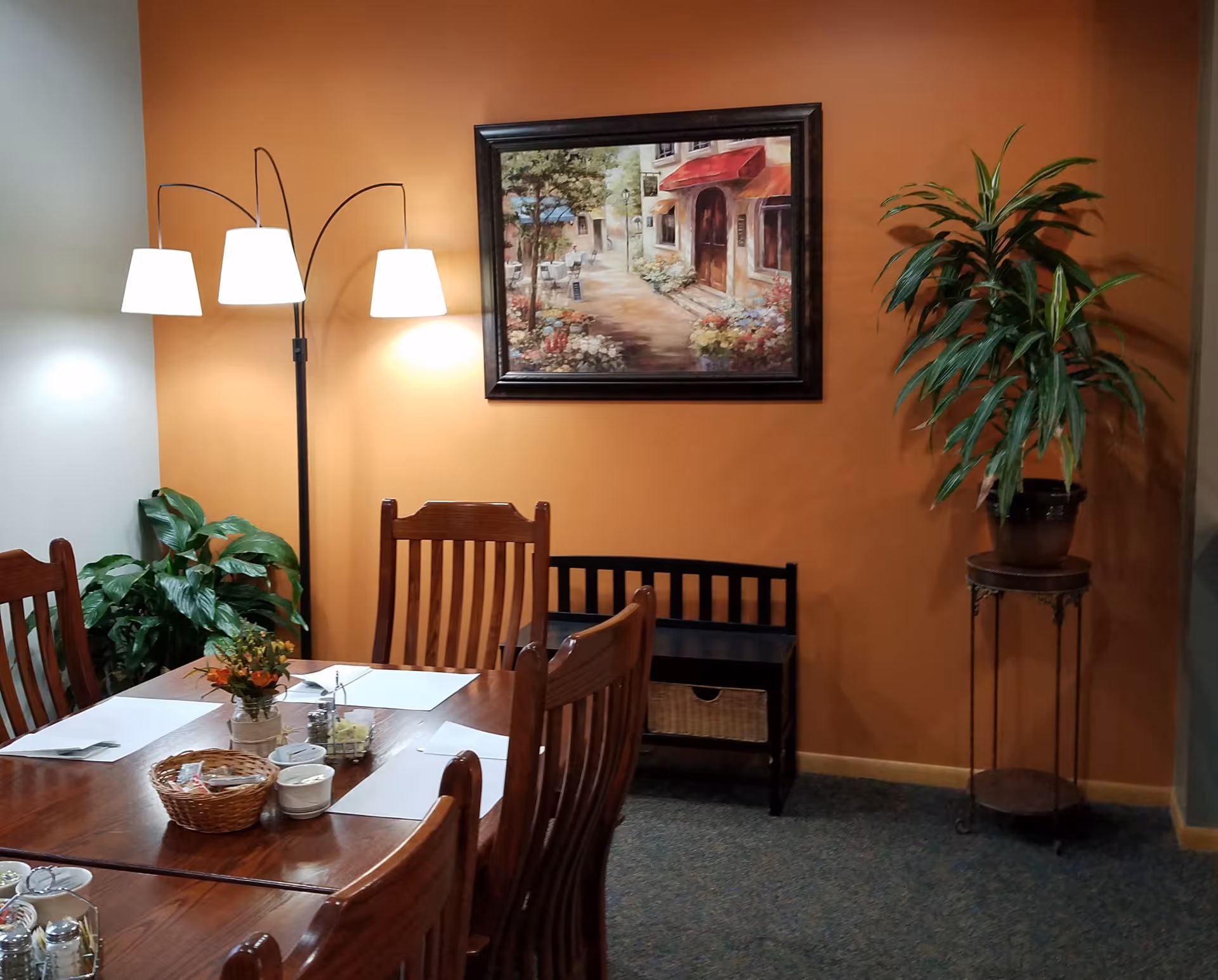 A dining area with a wooden table and chairs set against an orange wall. The table has placemats, a small basket, condiments, and a small flower arrangement. There is a three-lamp floor light, a framed painting of a street scene, and two potted plants on either side of the wall.