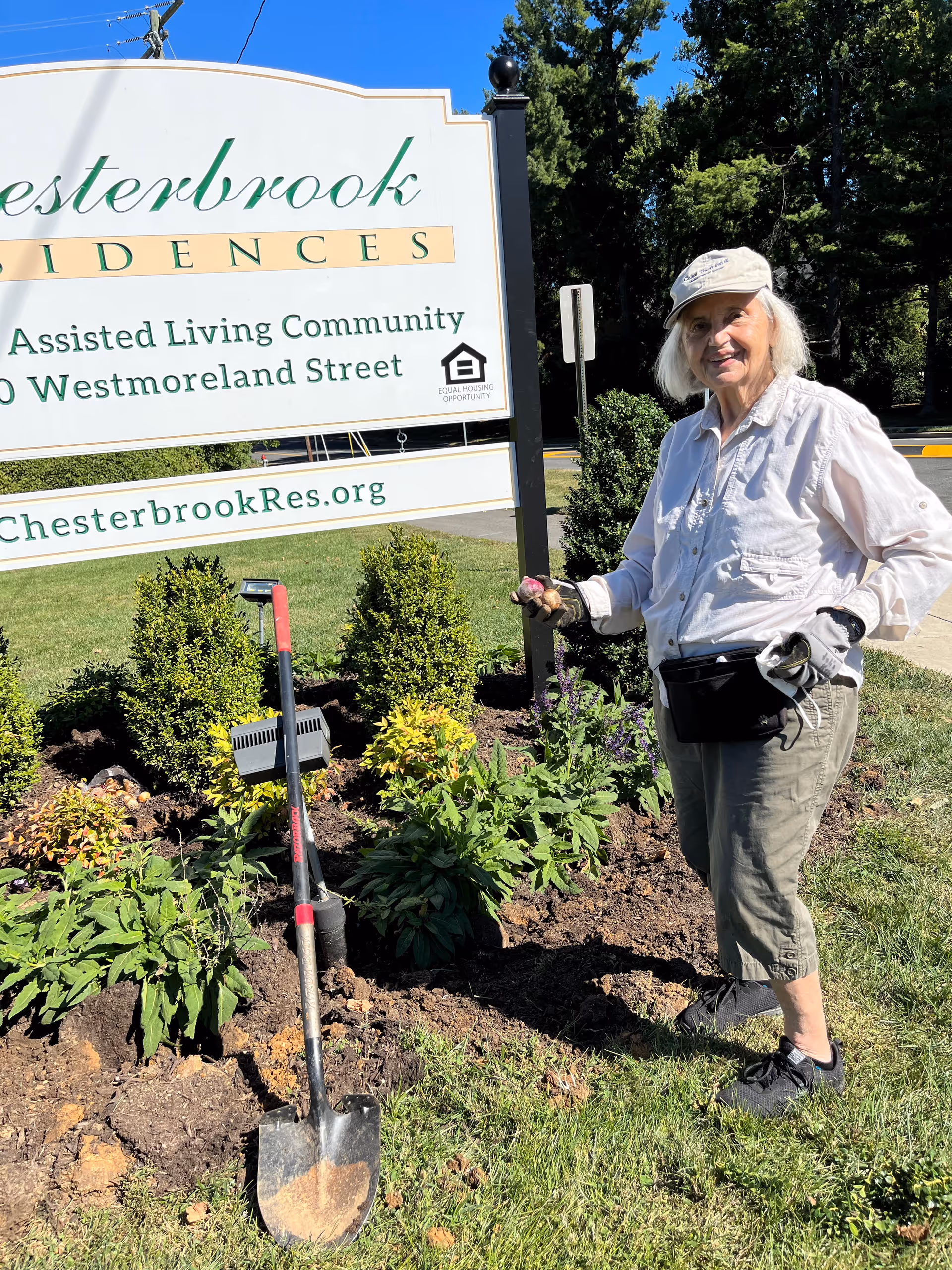 A woman wearing a hat stands next to a shovel and planted flower bed in front of a large Chesterbrook Residences sign.