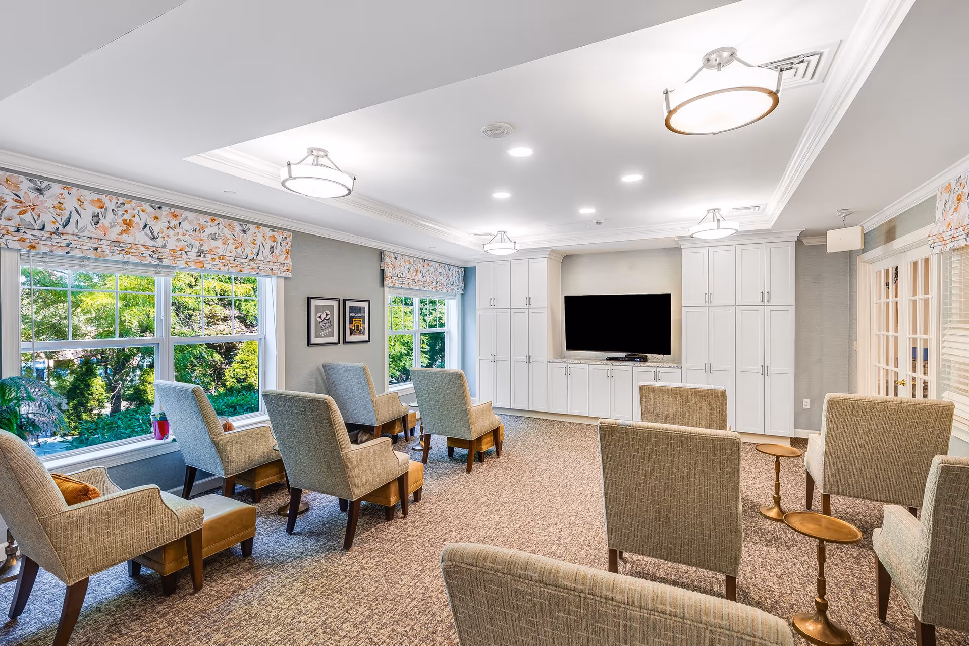 A bright and cozy common room with several upholstered armchairs arranged in rows facing a large flat-screen TV mounted on a wall with white cabinetry. The room has large windows with floral valances letting in natural light and a carpeted floor.