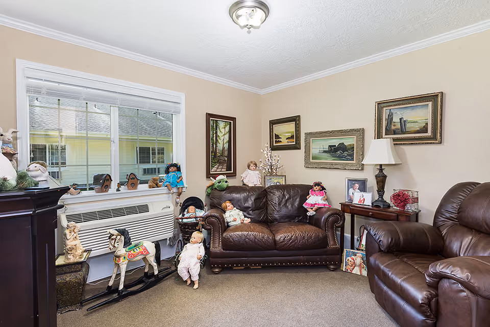 A cozy living room with beige walls and carpeted floor, featuring two brown leather sofas. The room is decorated with several dolls placed on the sofas, window sill, and floor. There are framed paintings on the walls, a table lamp on a side table, and a rocking horse near the window. A large window with blinds lets in natural light.