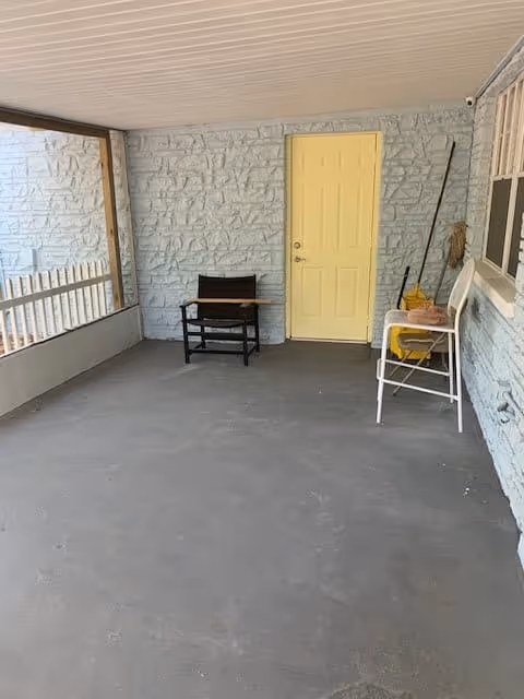 A covered porch area with a gray concrete floor and light blue stone walls. There is a yellow door in the center of the wall, a black chair with a wooden armrest to the left of the door, and a white bench with a yellow cushion and cleaning supplies to the right. The porch has a white ceiling and a railing on the left side.