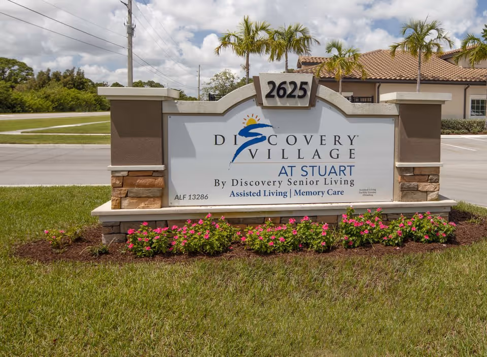 Outdoor view of a large sign for Discovery Village At Stuart, a senior living facility offering assisted living and memory care. The sign is set in a landscaped area with grass and pink flowers, with a building and palm trees visible in the background under a partly cloudy sky.