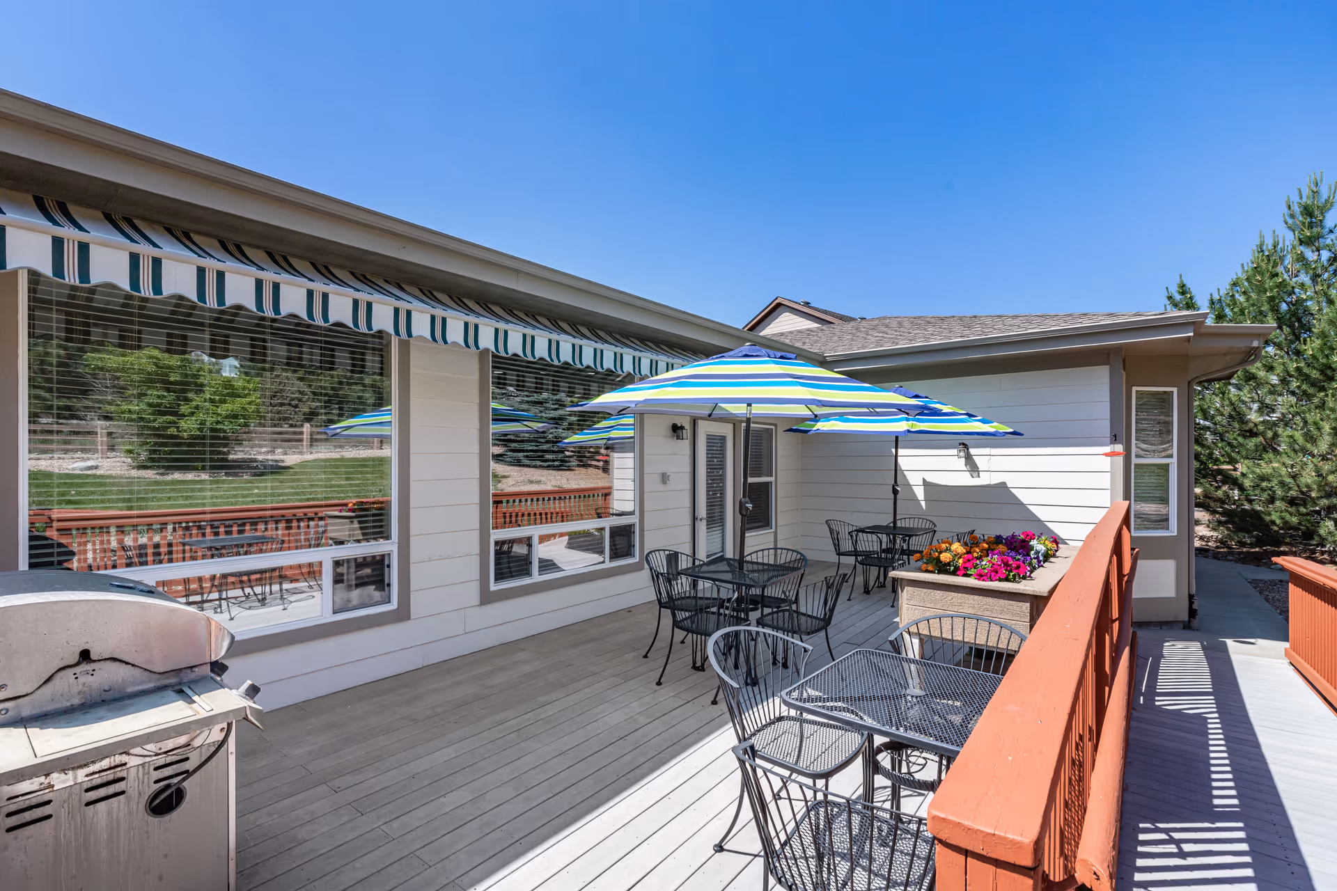 Outdoor patio area with metal tables and chairs, striped umbrellas providing shade, a flower planter, and a barbecue grill. The patio is attached to a building with large windows and an awning. The sky is clear and blue.