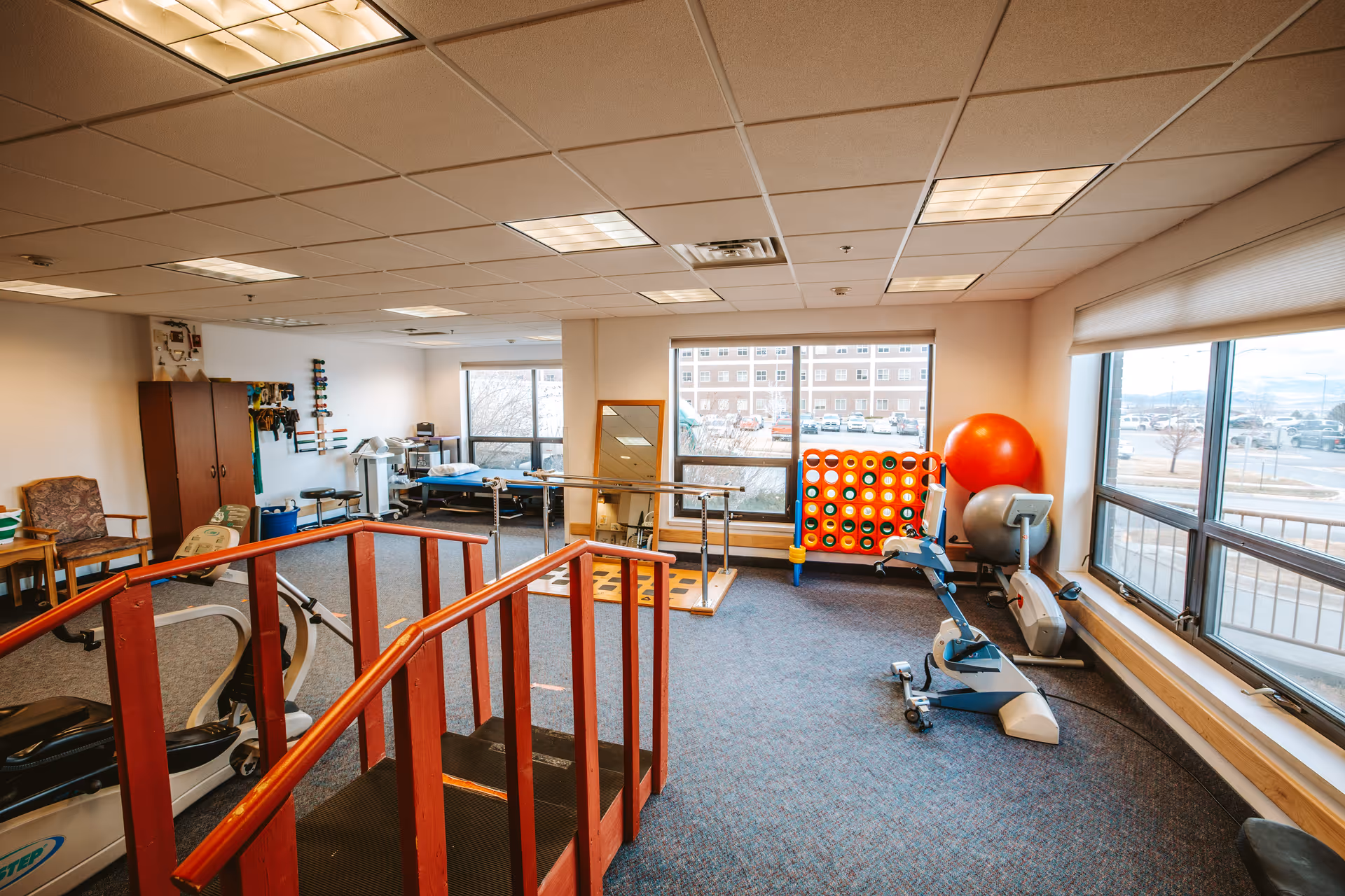 A rehabilitation therapy room with exercise equipment including a stationary bike, parallel bars for walking practice, a large Connect Four game, and therapy balls. The room has large windows letting in natural light and a carpeted floor.