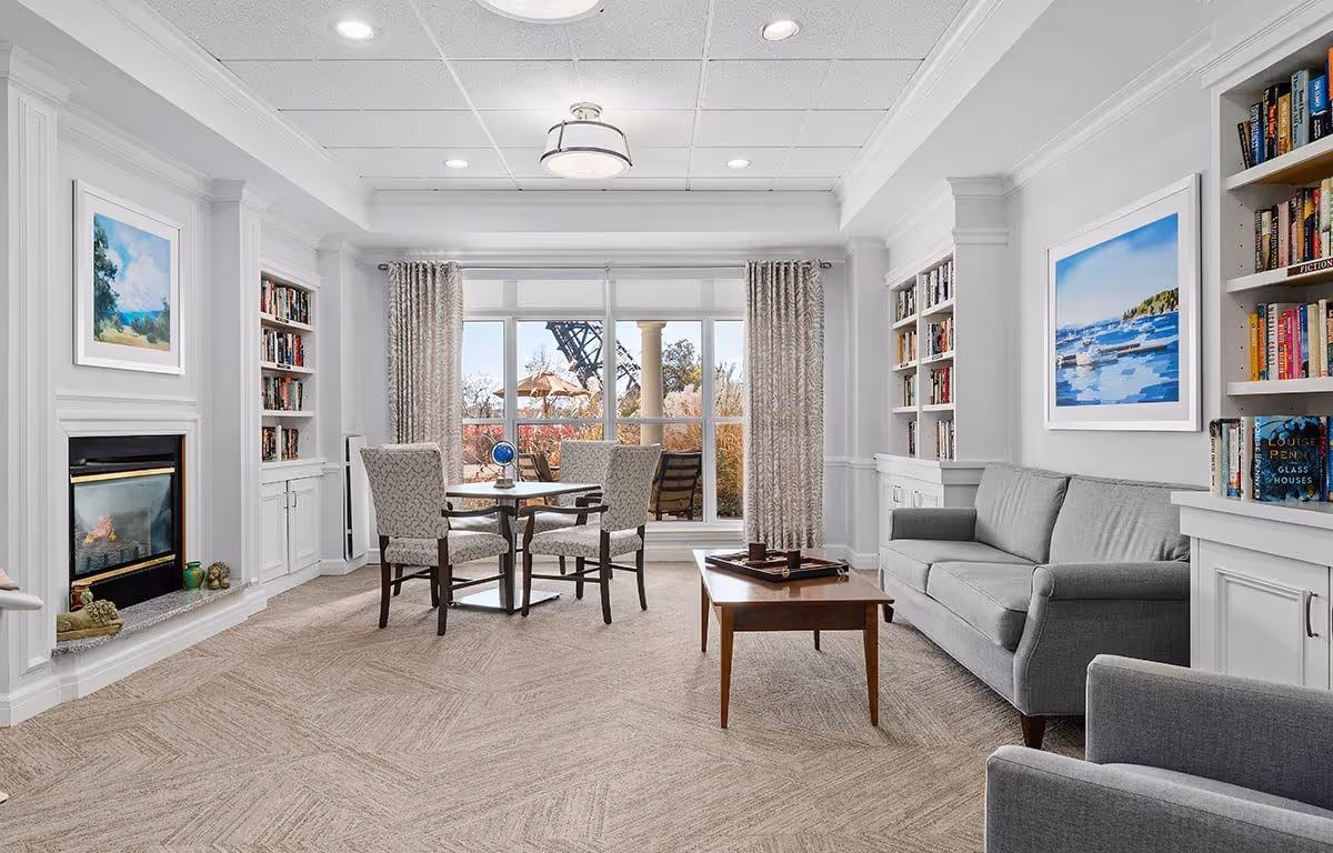 Bright common living room with sofas, chairs, a coffee table, built-in bookshelves, a fireplace and a table by large windows looking out to a patio.