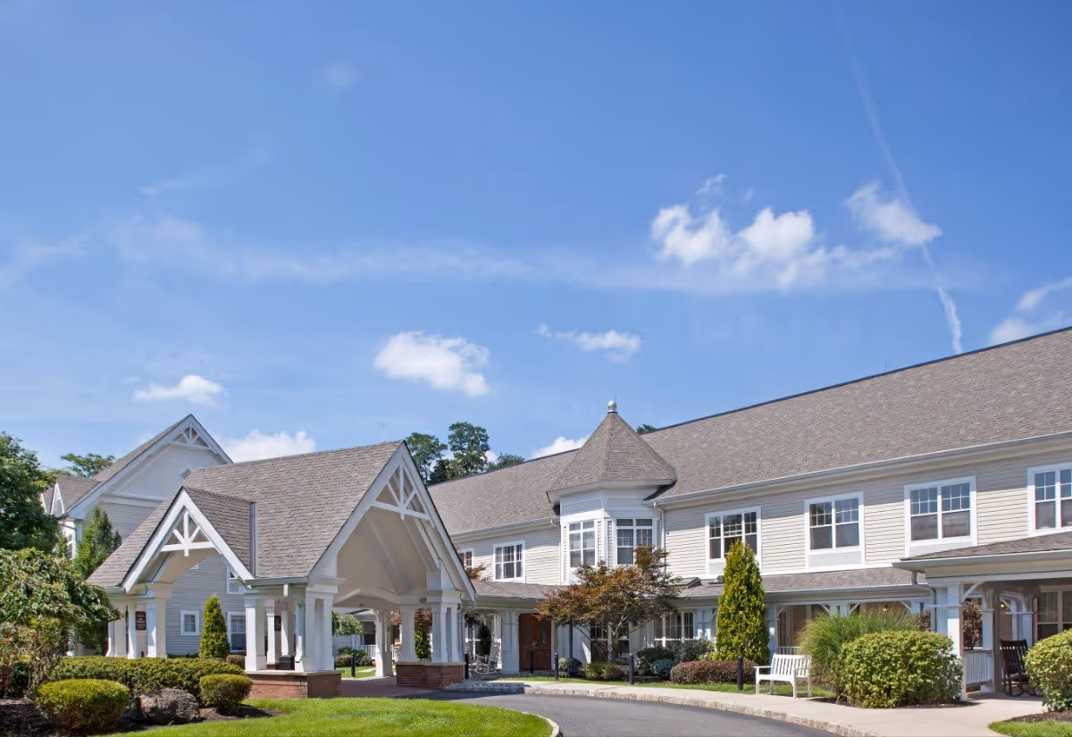 Exterior view of a senior living facility building with a covered entrance, manicured bushes, trees, and a clear blue sky with some clouds.