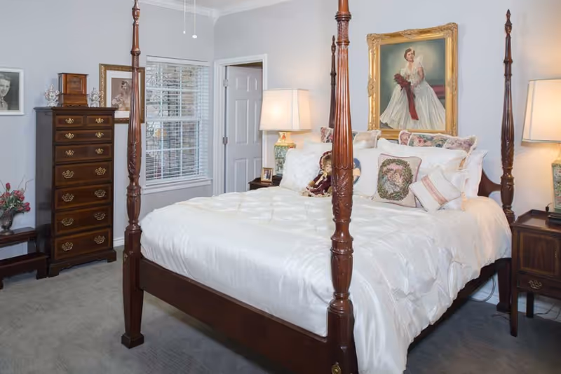 A traditional bedroom featuring a wooden four-poster bed with white bedding and decorative pillows. On either side of the bed are wooden nightstands with matching lamps. A tall wooden dresser stands against the wall near a window with white blinds. The walls are adorned with framed portraits, including a large painting of a woman in a white dress above the bed.