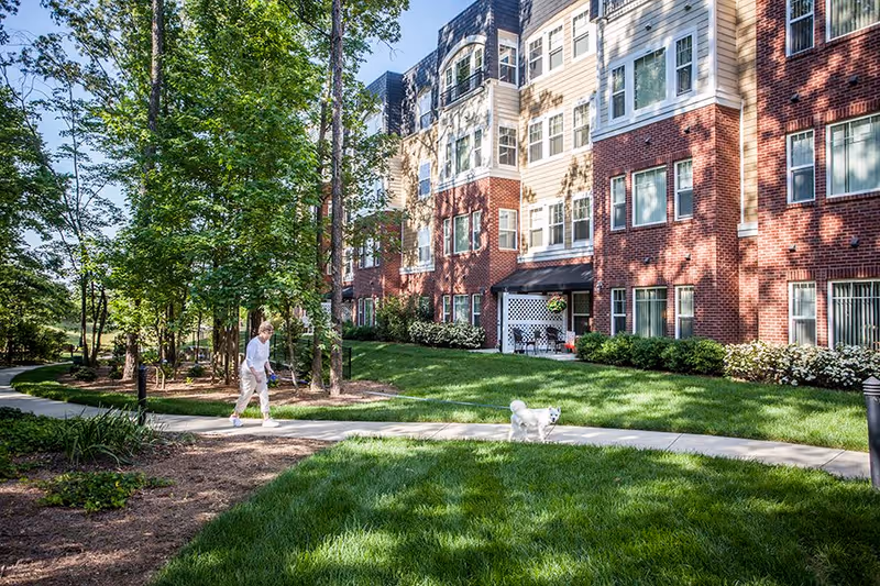 A senior woman walking a small white dog on a leash along a curved sidewalk in a landscaped garden area with green grass and trees, next to a multi-story brick and siding residential building with large windows and a small patio area.