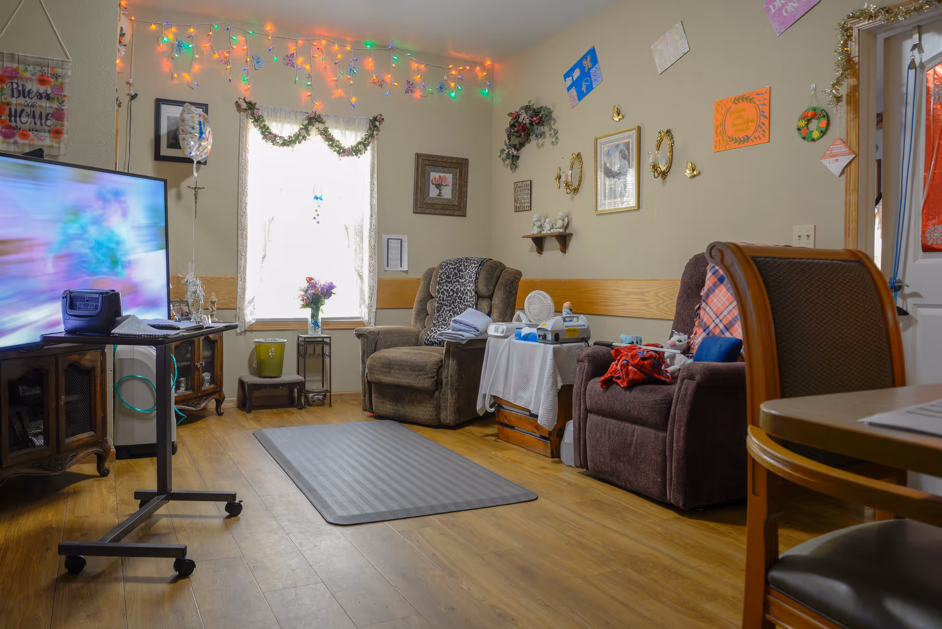 A cozy living room with two recliner chairs, a small table with medical equipment, a TV on a stand, and festive string lights hanging near the window. The walls are decorated with framed pictures, wreaths, and colorful paper decorations. The floor is wooden with a gray rug in the center.