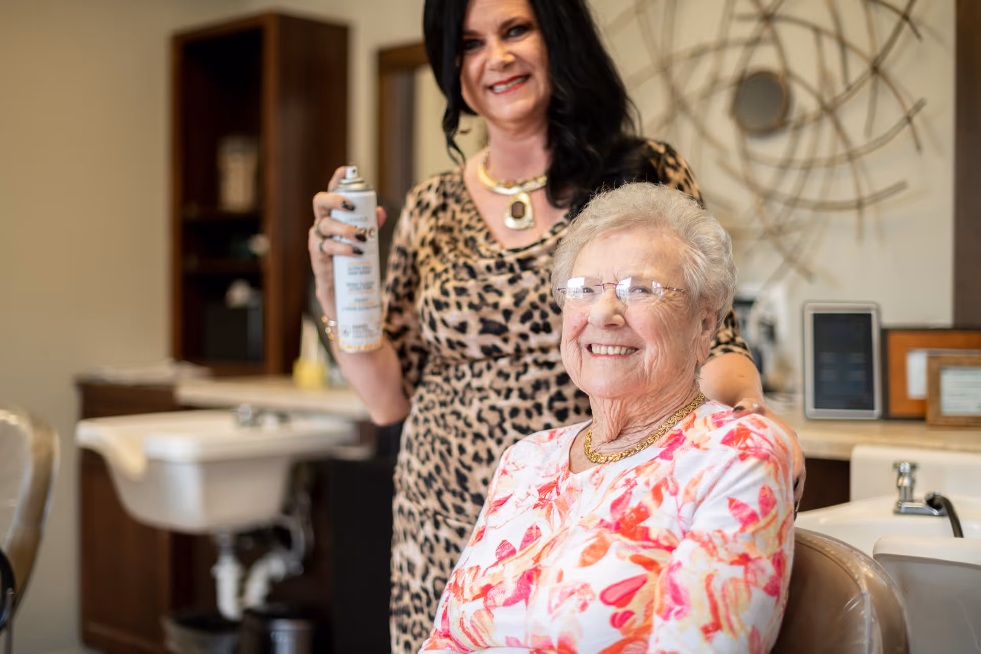 An elderly woman with glasses and a floral top is sitting in a salon chair, smiling. Behind her stands a hairstylist holding a can of spray, both inside a salon area with sinks and shelves in the background.