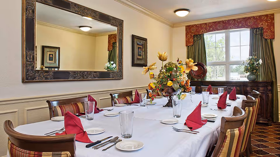 A formal dining room with a long table covered with a white tablecloth, set with plates, glasses, silverware, and red folded napkins. The room features wooden chairs with striped cushions, a large decorative mirror on one wall, a window with green curtains and a red valance, and floral centerpieces on the table and a sideboard.