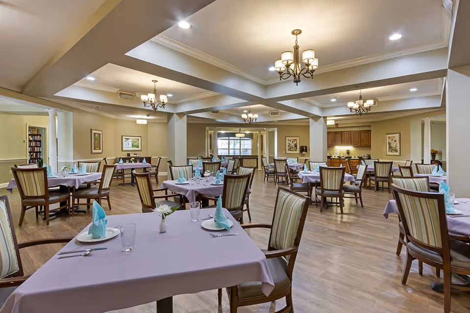 A spacious dining room with multiple tables covered in light purple tablecloths, each set with plates, glasses, silverware, and folded blue napkins. The room features wooden chairs with striped upholstery, chandeliers hanging from a coffered ceiling, and framed artwork on the beige walls. There is a kitchen area visible in the background.