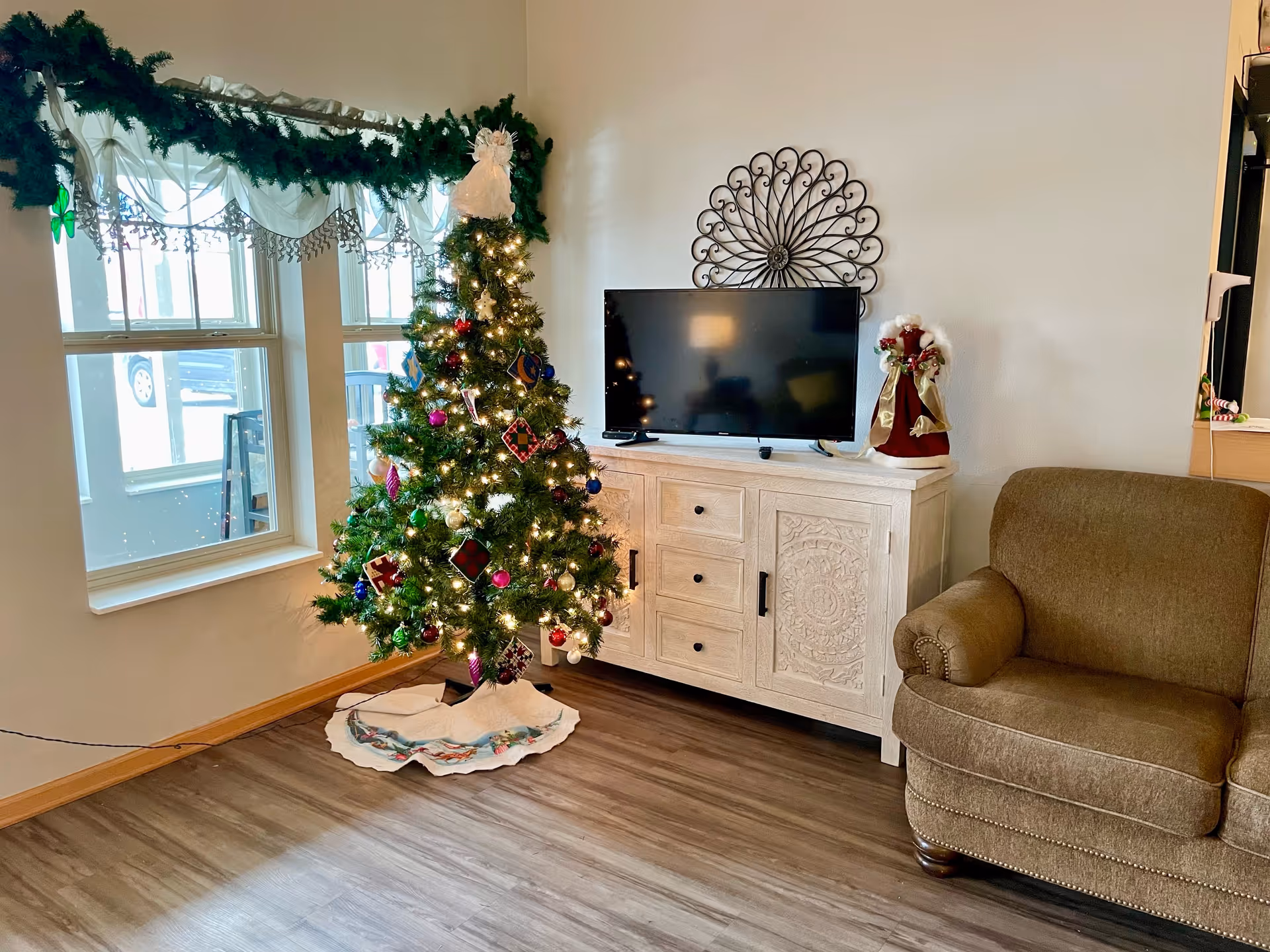 A cozy living room corner decorated for Christmas with a lit Christmas tree adorned with ornaments and a white tree skirt. Next to the tree is a white wooden cabinet with intricate carvings, holding a flat-screen TV and a Santa Claus figurine. A brown upholstered armchair is positioned to the right, and a window with white curtains and green garland is on the left.