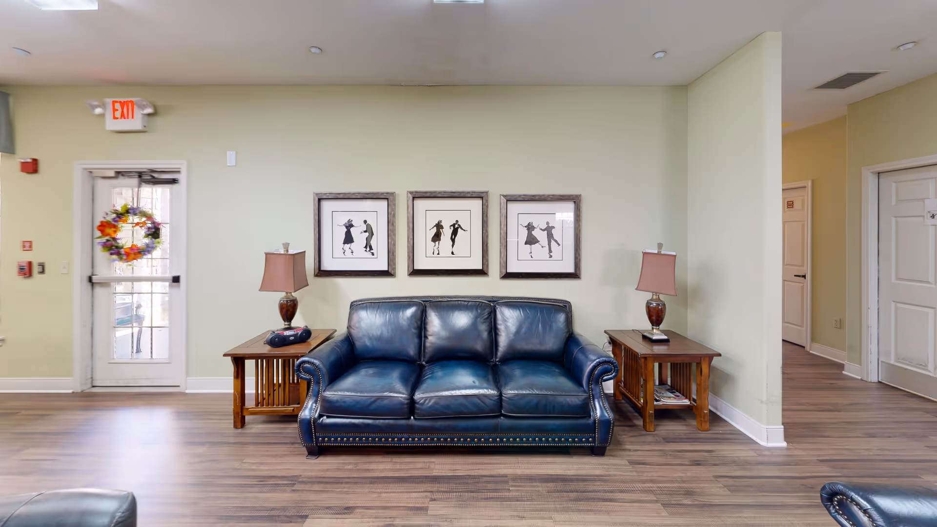 A cozy seating area in a senior living facility with a dark blue leather couch centered against a light green wall. On either side of the couch are wooden side tables with matching lamps. Above the couch are three framed black and white silhouette art pieces depicting dancing figures. To the left is a glass door decorated with a colorful wreath, and to the right are doorways leading to other rooms. The floor is wood laminate.