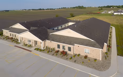 Aerial view of a single-story brick building with a dark shingled roof, surrounded by a parking lot and open fields. The building has multiple windows and landscaped areas with small bushes and flowers along the perimeter.