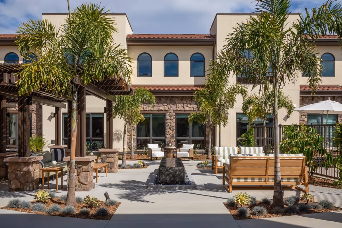 Outdoor courtyard area at Westmont of Carmel Valley featuring palm trees, stone and stucco building facade, seating areas with cushioned chairs and sofas, a central water feature, and shaded pergolas under a partly cloudy sky.