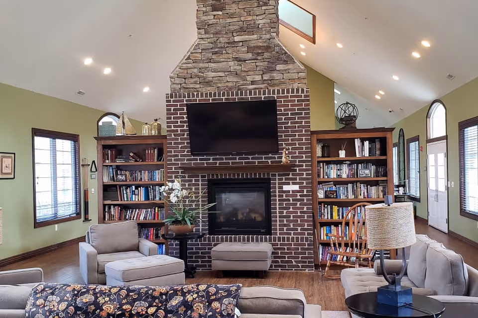 A cozy living room with a high vaulted ceiling featuring a central brick and stone fireplace with a mounted flat-screen TV above it. On either side of the fireplace are wooden bookshelves filled with books and decorative items. The room has green walls, multiple windows with blinds, and comfortable seating including armchairs, a sofa, and an ottoman. A table lamp and a floral arrangement add to the warm ambiance.