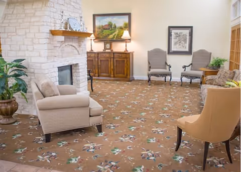 A cozy living room area with beige armchairs and a sofa arranged around a stone fireplace. The room has a floral patterned carpet, a wooden cabinet with two lamps and a painting above it, and framed artwork on the wall. There is also a potted plant in the corner.