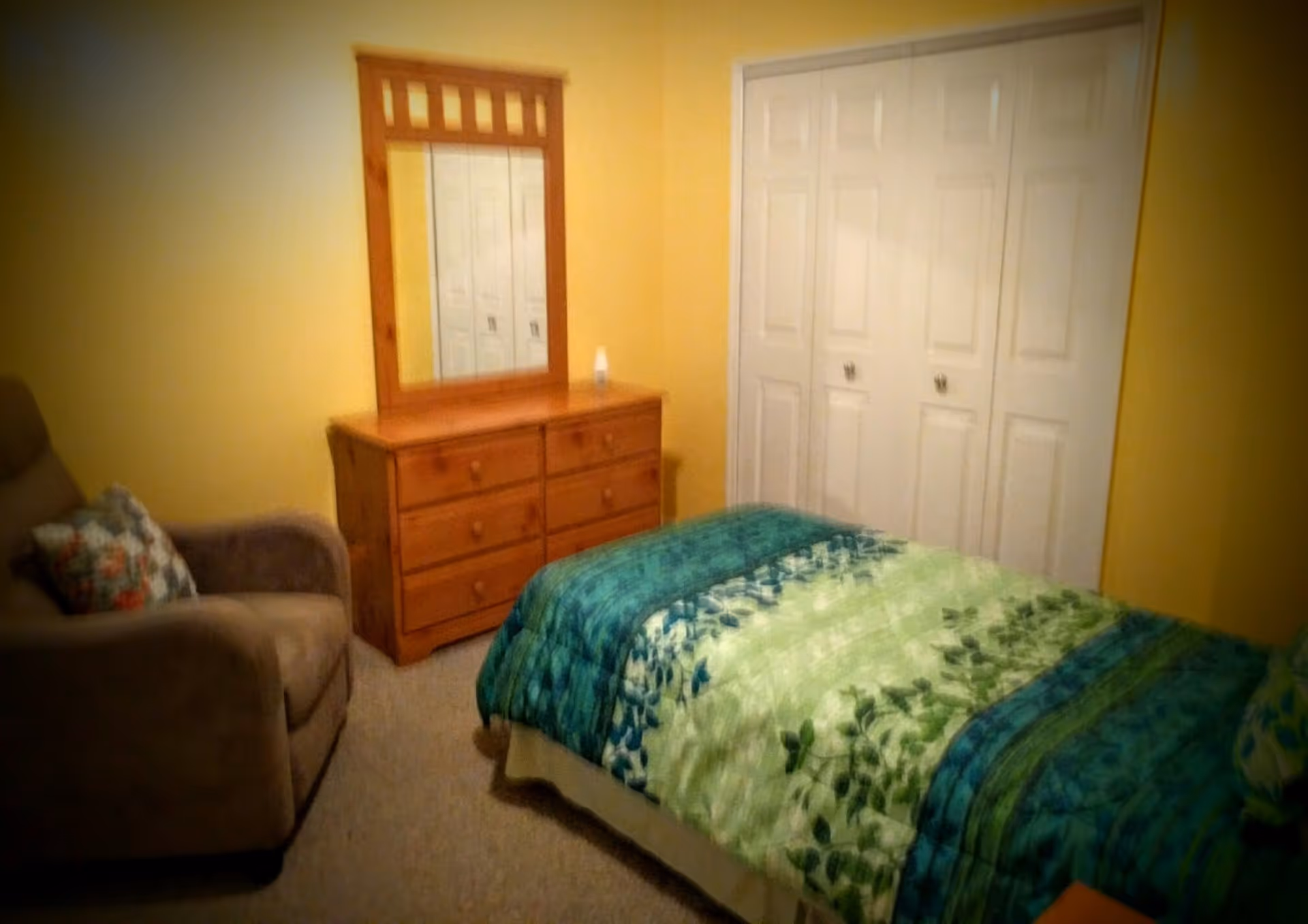 A cozy bedroom with yellow walls featuring a bed with a green and blue patterned comforter, a wooden dresser with a mirror, a beige armchair with a floral pillow, and white double closet doors.