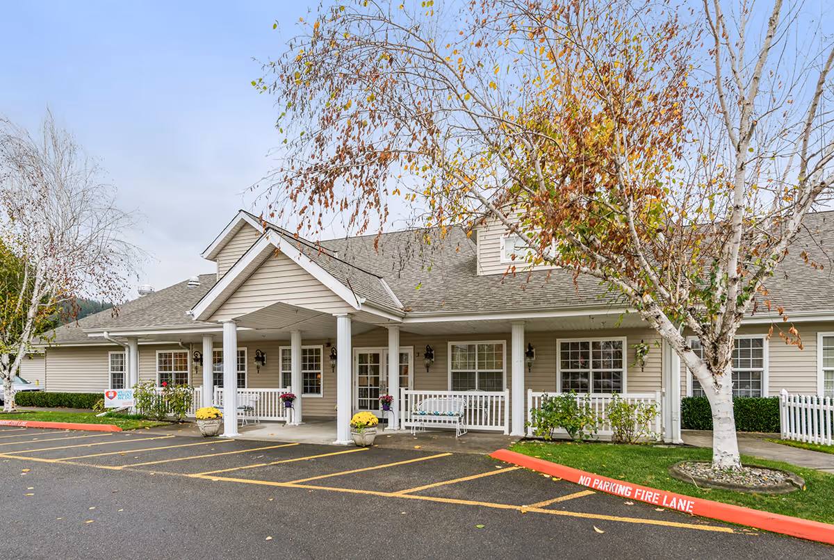 Exterior view of Avista Senior Living Sumner building with beige siding, white columns, and a covered entrance. There are trees with autumn leaves and a parking area with marked fire lane in front.