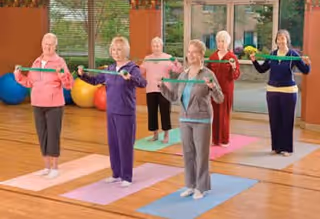 Five elderly women standing on yoga mats in a fitness room, holding resistance bands stretched in front of them as part of an exercise routine. The room has large windows, wooden floors, and colorful exercise balls in the background.