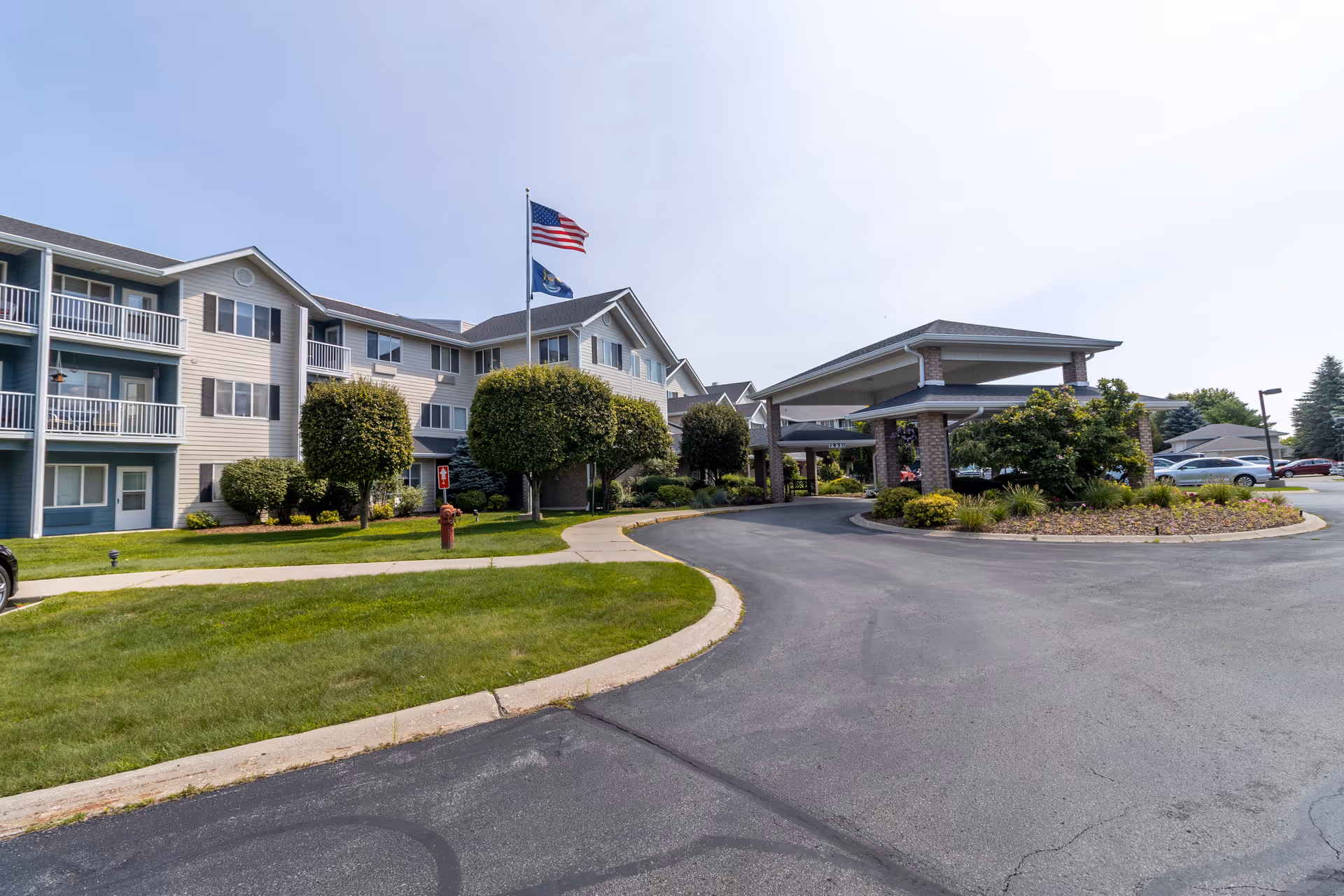 Exterior view of Independence Village of Traverse City, showing a multi-story building with balconies, a covered entrance driveway, well-maintained landscaping, and two flagpoles with the American flag and another flag.