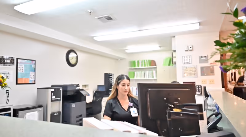 A woman in a black uniform working at a reception desk with a computer monitor in an office setting. The background shows office equipment, filing shelves with green folders, a clock on the wall, and some posters.