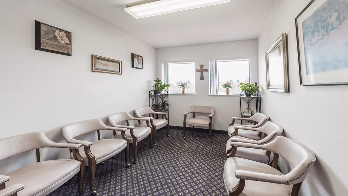 A small waiting or meeting room with eight beige cushioned chairs arranged along the walls. The room has two windows with potted plants on black metal shelves beneath them. The walls are decorated with framed artwork and a wooden cross between the windows. The carpet is dark with a patterned design, and the ceiling has a fluorescent light fixture.