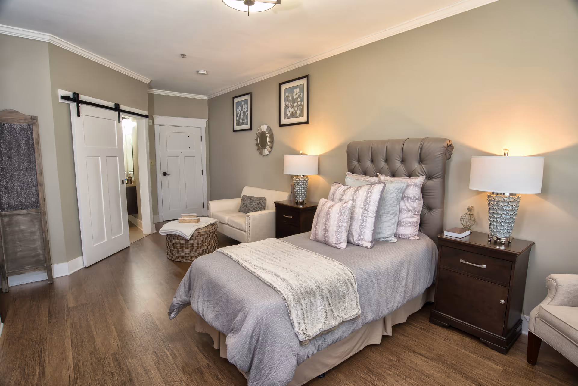 Sunlit bedroom with a tufted single bed, two nightstands with lamps, an armchair, and a sliding barn door leading to a bathroom.