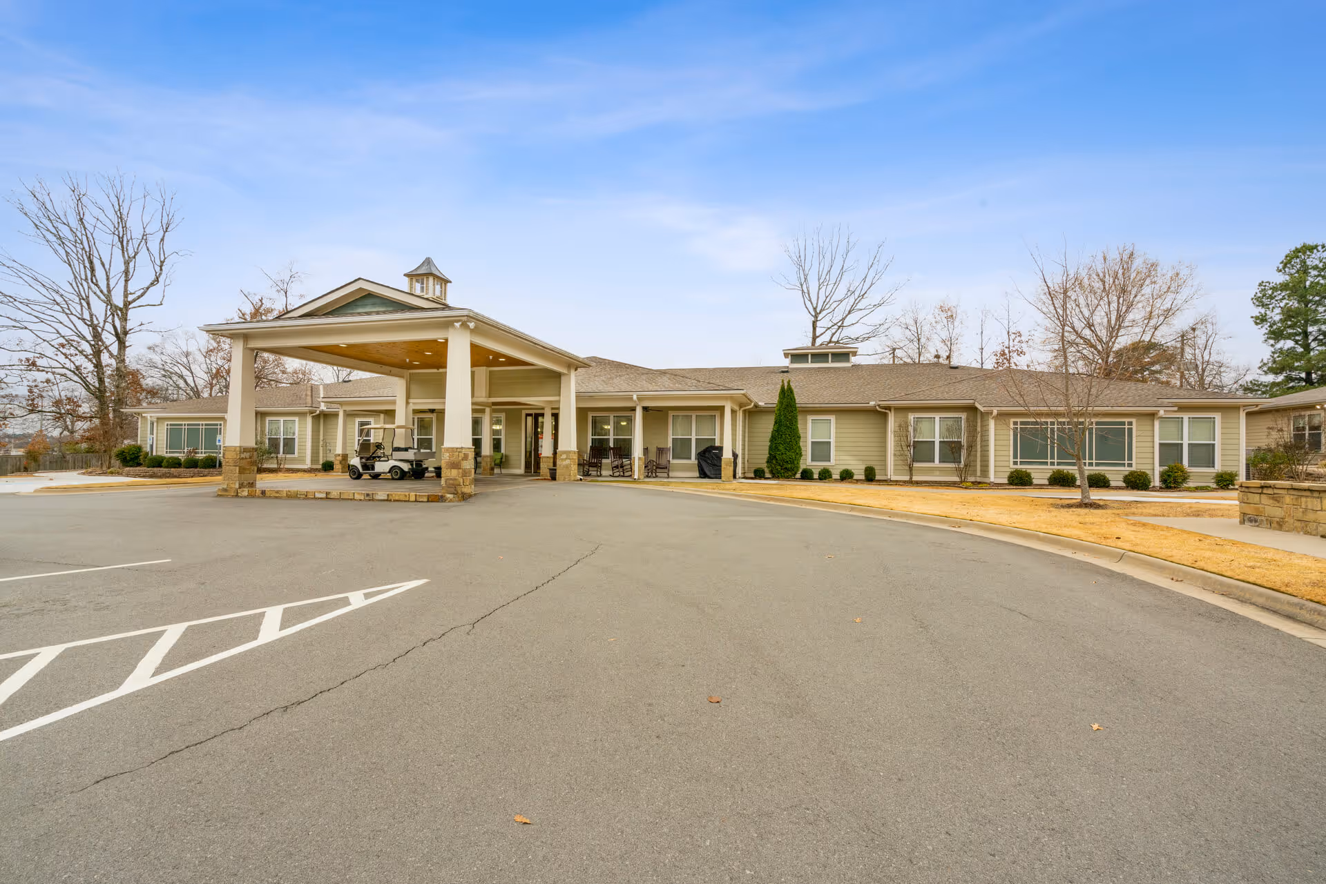 Front exterior view of a single-story senior living facility building with a covered entrance, a golf cart parked under the canopy, leafless trees, and a clear blue sky.