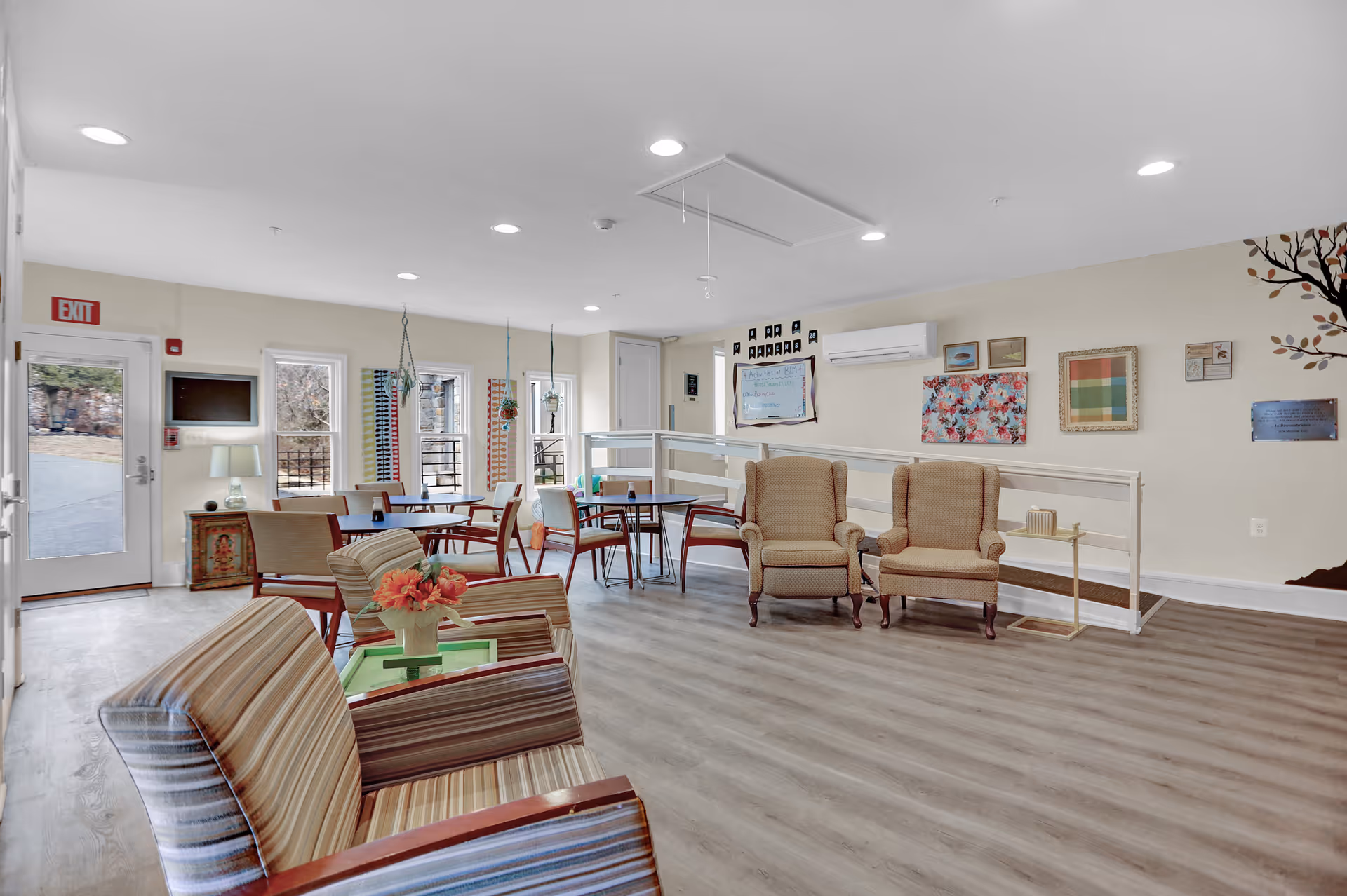 A bright and spacious common area in an assisted living facility featuring several armchairs and tables with chairs arranged for socializing or activities. The room has light-colored walls decorated with framed artwork and a wall-mounted air conditioning unit. Large windows and a glass door provide natural light and a view of the outdoors. The floor is covered with wood-style flooring.