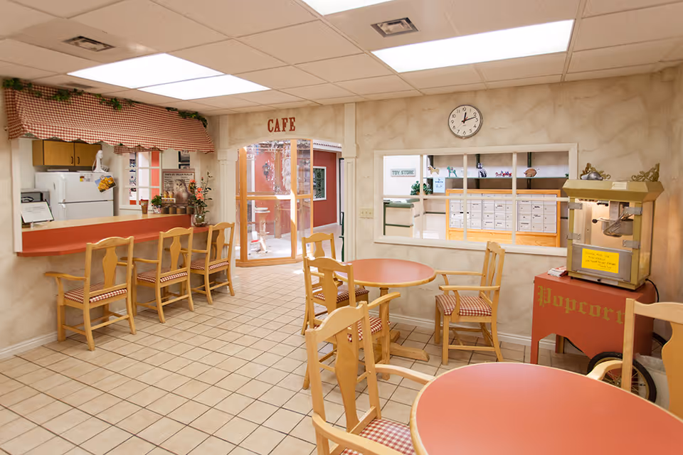 Interior view of a cozy cafe area in a senior living facility named Quail Ridge, featuring round tables with wooden chairs that have checkered cushions, a popcorn machine on a red cart, a counter with bar stools, a clock on the wall, and a window looking into another room labeled 'Toy Store'.