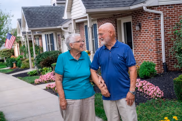 An elderly couple holding hands and smiling at each other while standing on a sidewalk in front of a row of brick residential buildings with well-maintained gardens and an American flag.