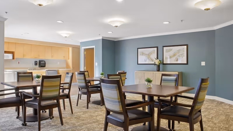 A bright dining room in Aspenwood Senior Living Community featuring several wooden tables with striped cushioned chairs. The room has light blue walls adorned with two framed abstract artworks and a sideboard with a vase of flowers. The background shows a kitchenette area with light wood cabinets, a microwave, and a sink.