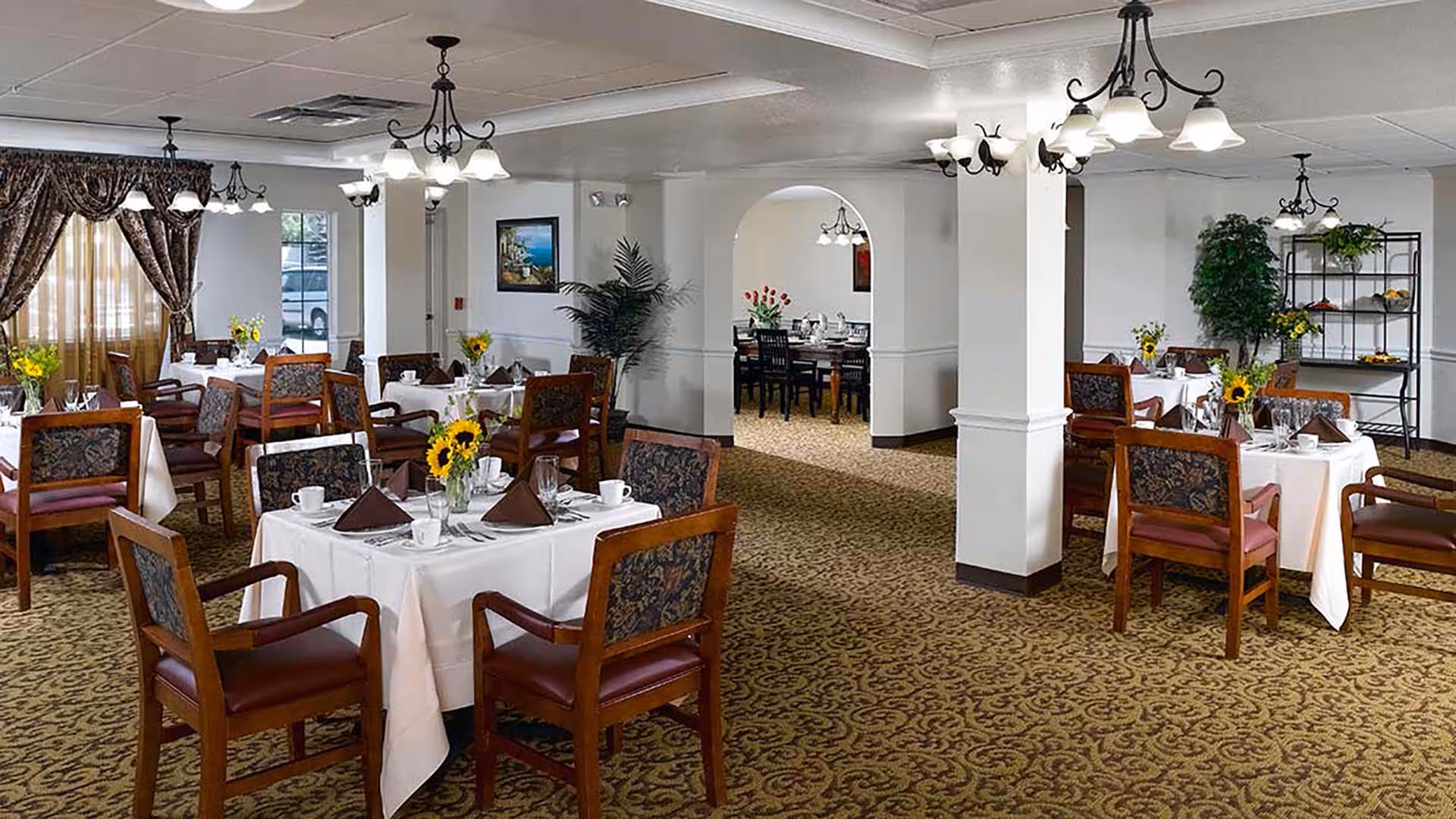 A spacious dining room with multiple tables covered in white tablecloths, each set with cups, napkins, and small flower arrangements. The room features patterned carpet, wooden chairs with upholstered backs and seats, chandeliers hanging from the ceiling, and large windows with heavy drapes allowing natural light to enter. There are plants and framed artwork on the walls, and an archway leads to another dining area in the background.