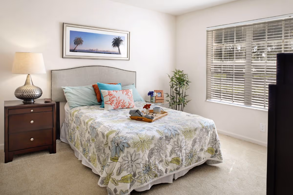 A bright and cozy bedroom featuring a bed with a floral patterned bedspread and multiple pillows. There is a wooden nightstand with a decorative lamp on the left side of the bed. A framed picture of palm trees and a city skyline hangs above the headboard. To the right, a window with blinds lets in natural light, and a small round table with a plant and photo frame is placed near the corner.