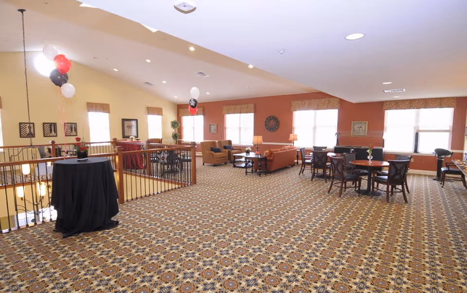 Spacious senior living common area with patterned carpet, seating groups, small dining tables, and a few balloons.