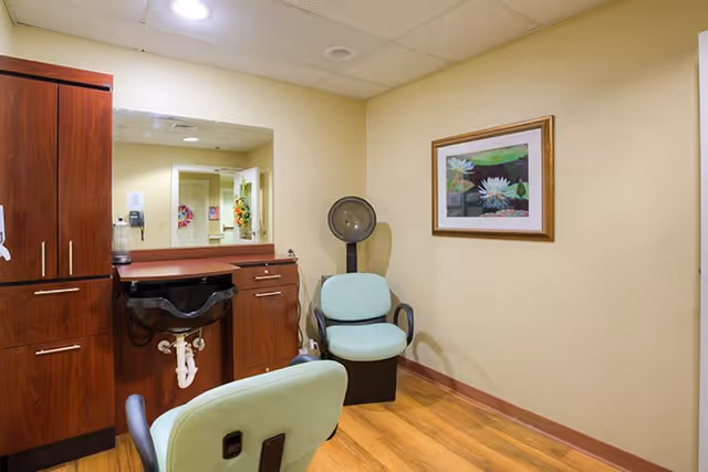 Small salon/styling room with two mint-green salon chairs, a hooded hair dryer, a sink and wood cabinets beneath a mirror.