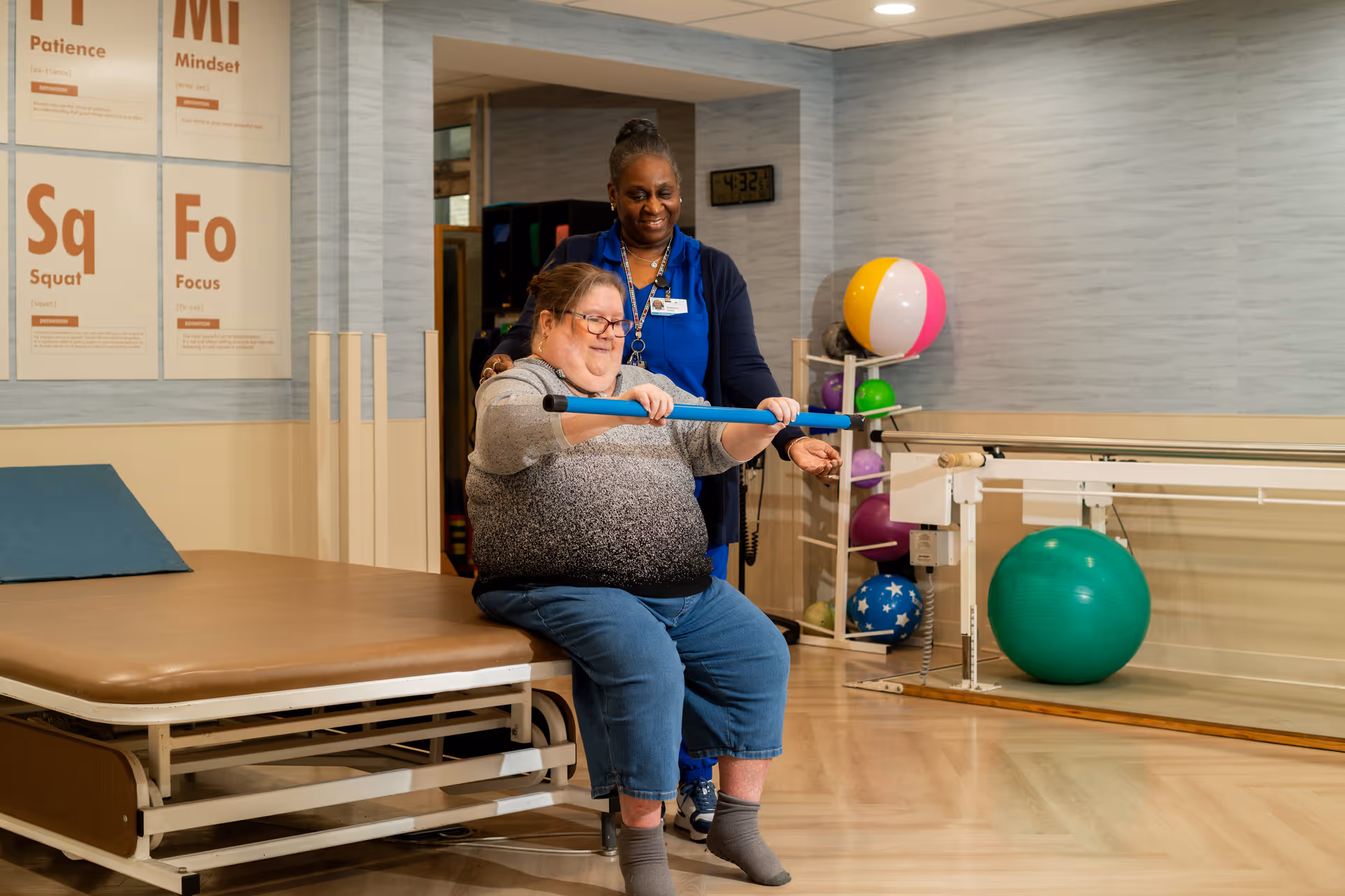 A woman is seated on a therapy table holding a blue exercise bar while a healthcare worker stands behind her providing support in a rehabilitation room equipped with exercise balls and parallel bars.