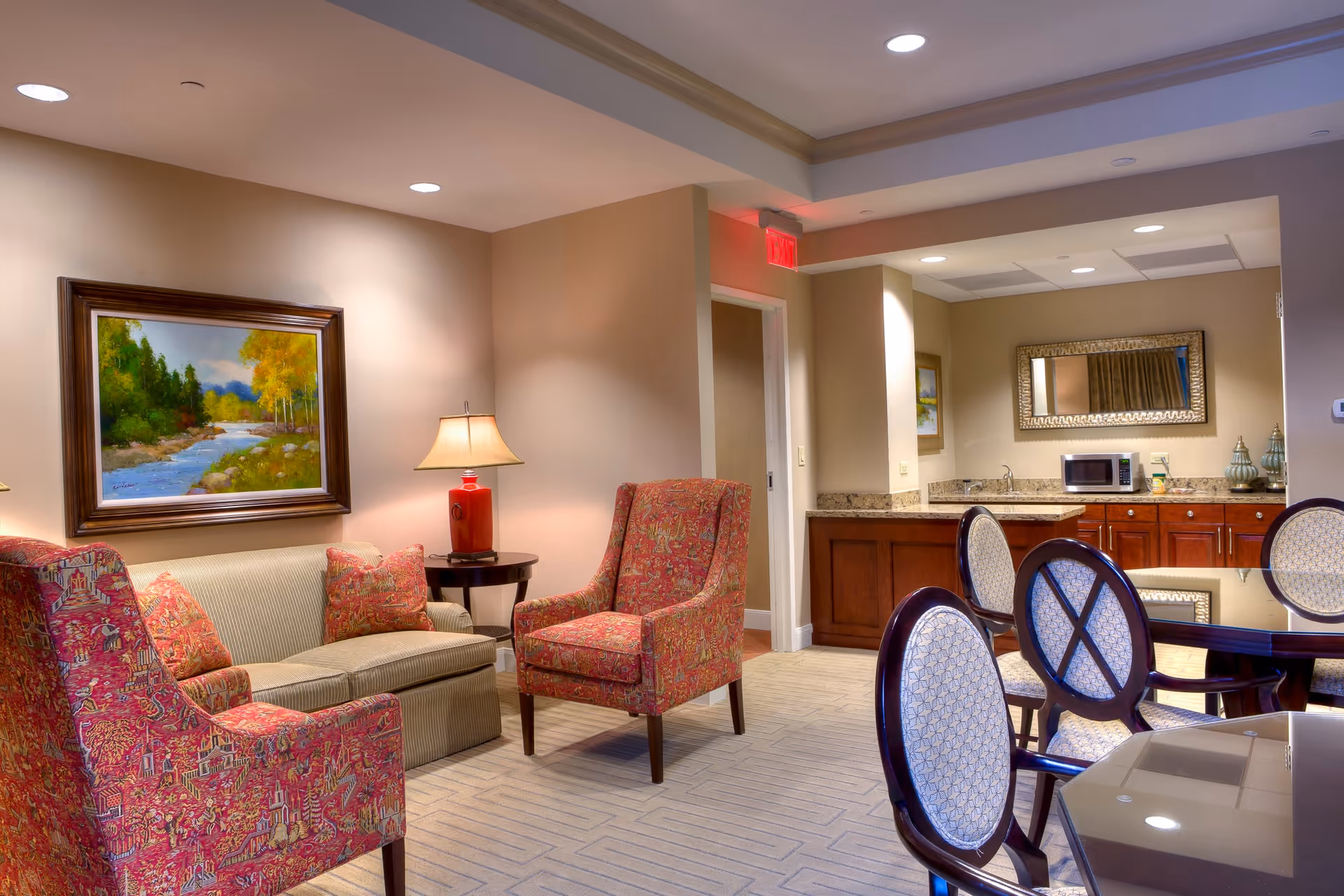 A cozy interior room featuring a seating area with a beige sofa and two red patterned armchairs around a small round table with a lamp. On the wall above the sofa is a framed painting of a river and trees. To the right, there is a dining table with four chairs and a kitchenette area with wooden cabinets, a microwave, a sink, and a large decorative mirror above the counter.