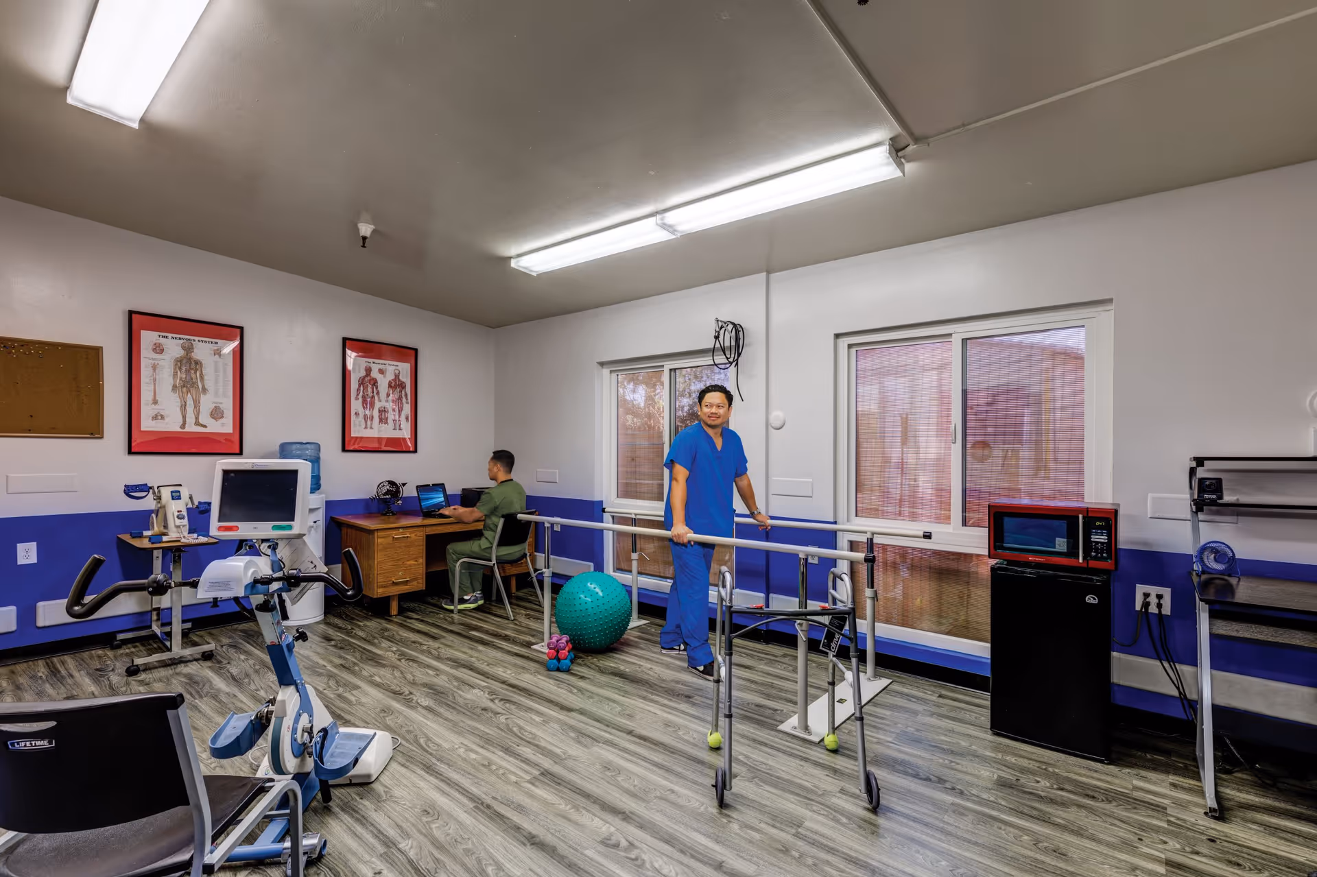 A rehabilitation or physical therapy room with two healthcare workers. One is sitting at a desk using a laptop, and the other is standing near parallel bars used for walking exercises. The room has medical posters on the wall, exercise equipment including a stationary bike, a large green exercise ball, and small dumbbells. There is a microwave and a small refrigerator against the wall, and the floor is covered with wood-patterned vinyl.