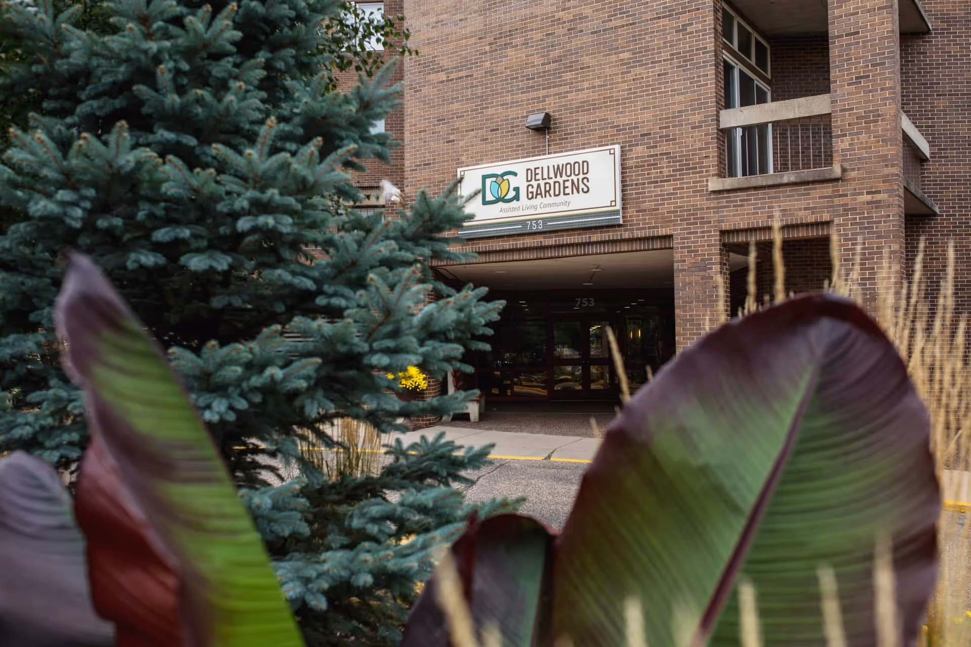 Front entrance of Dellwood Gardens assisted living community with a sign above the entry and plants in the foreground.