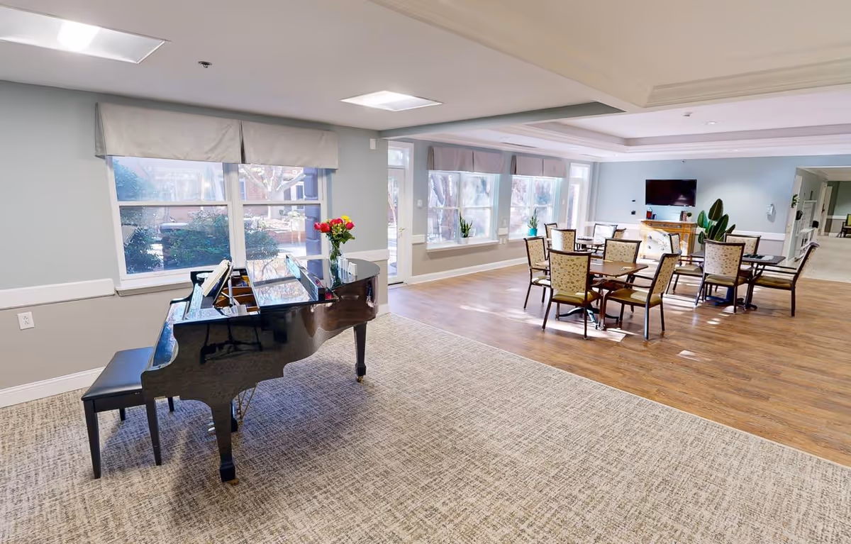 A spacious common area in a senior living facility featuring a black grand piano with a vase of flowers on top, several tables with chairs arranged for socializing or dining, large windows letting in natural light, and a wall-mounted television in the background.