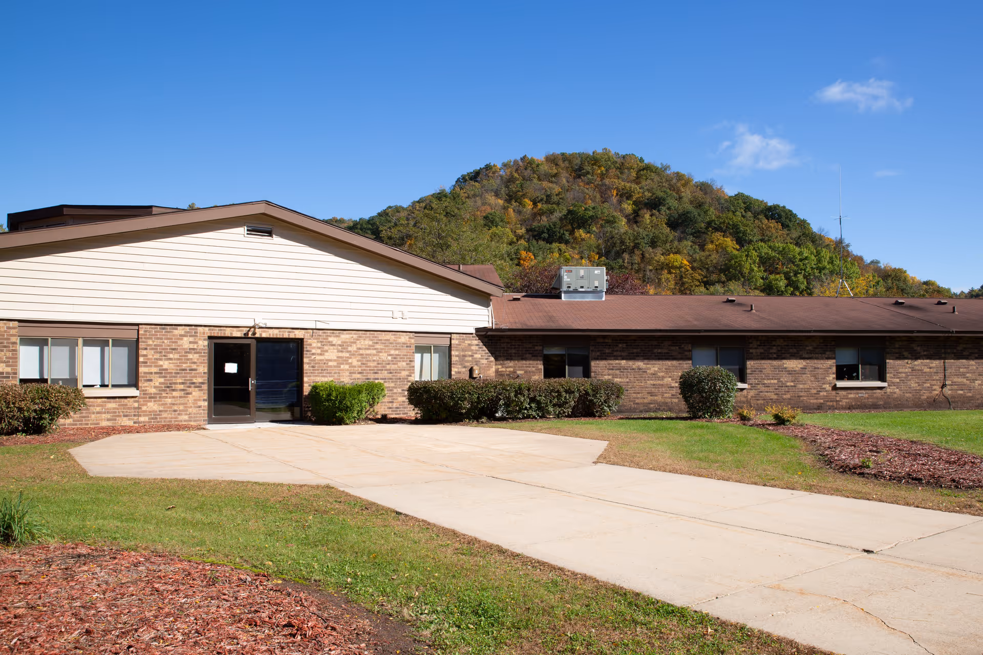 Low single-story brick building entrance with a paved driveway, shrubs, and a tree-covered hill behind under a clear blue sky.