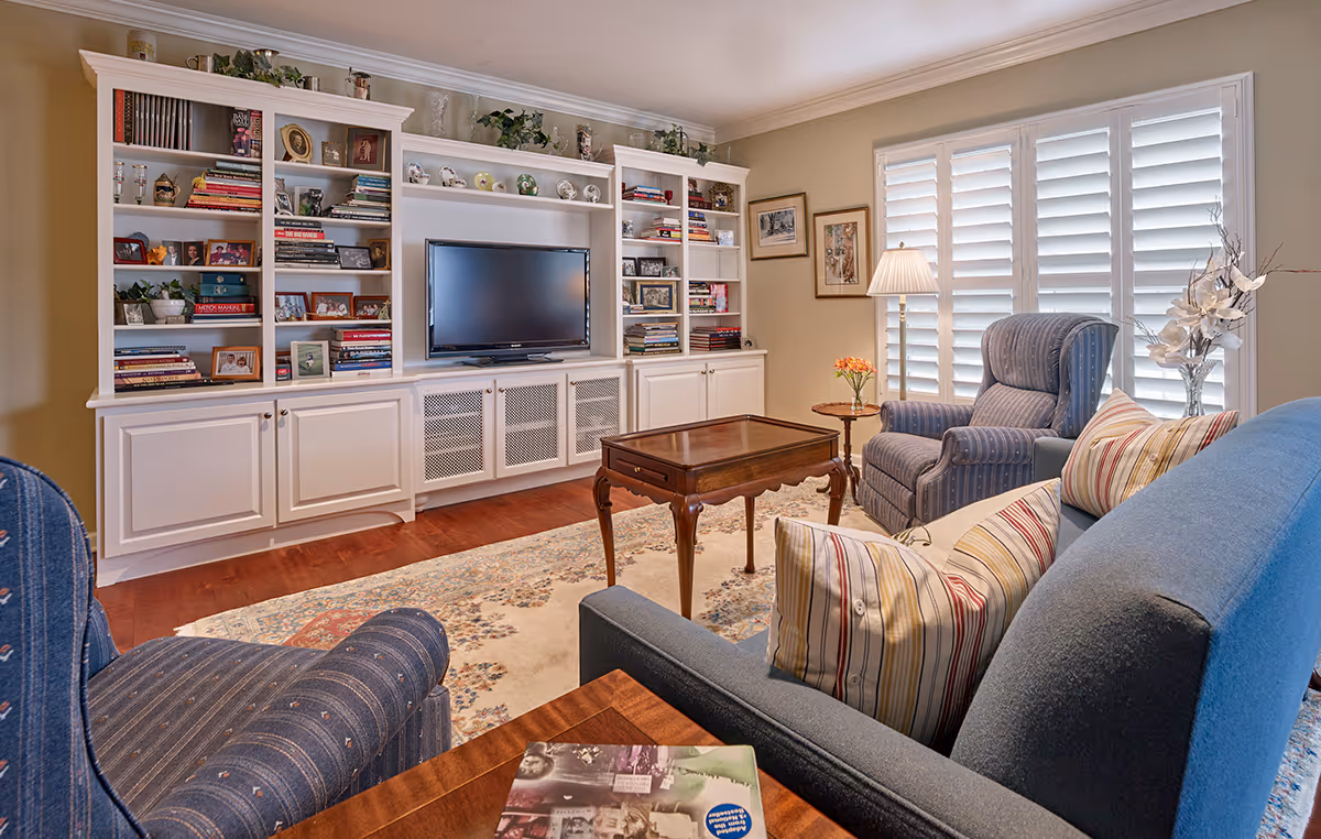 Cozy living room with built-in white bookshelves and TV, upholstered armchairs and a sofa around a wooden coffee table and shuttered windows.